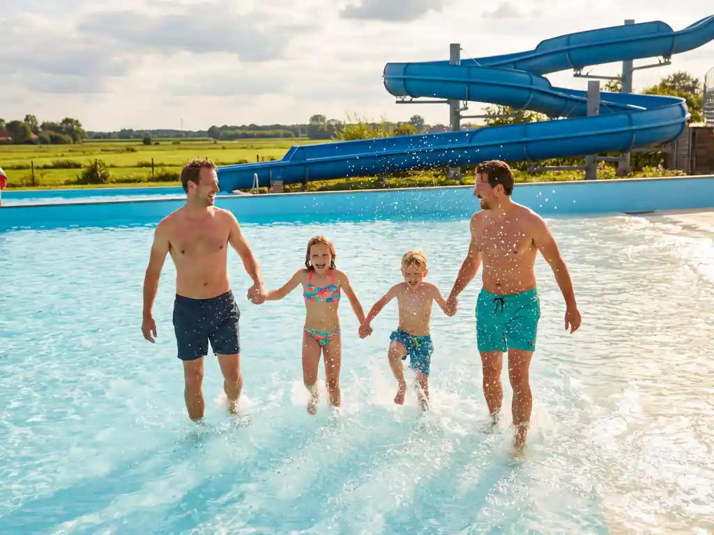 Familie met kinderen springt door golven in zwembad met blauwe glijbanen en groen Nederlands landschap op zonnige middag
