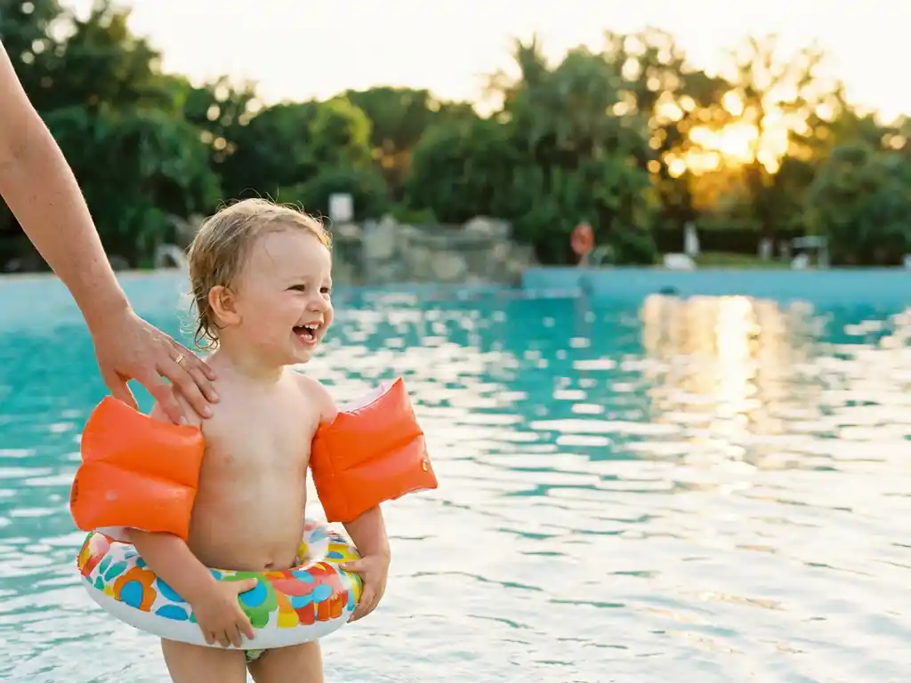 Peuter met oranje zwembandjes houdt opblaasbare ring vast aan rand van golfslagbad, ouderhand op schouder, gouden zonlicht op turquoise water