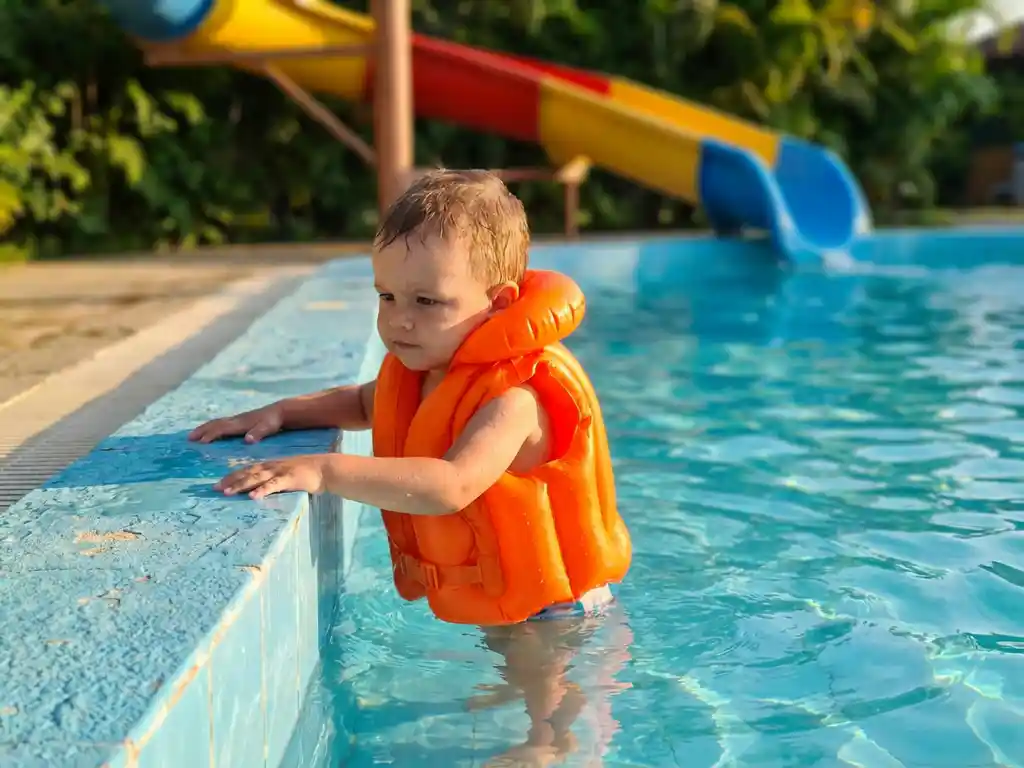 Kind met oranje zwemvest houdt zich vast aan de rand van een turquoise zwembad, gouden zonlicht schittert op het water