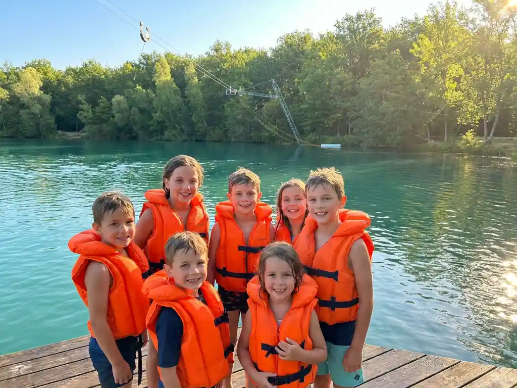 Groep kinderen in oranje zwemvesten op houten steiger bij kabelbaan, turquoise meer en groen bos op zonnige ochtend