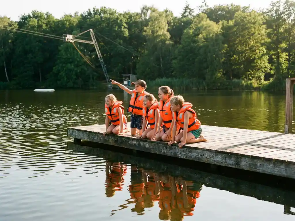 Enthousiaste kinderen in oranje zwemvesten op een houten steiger bij een meer, omringd door groen bos in warm middaglicht