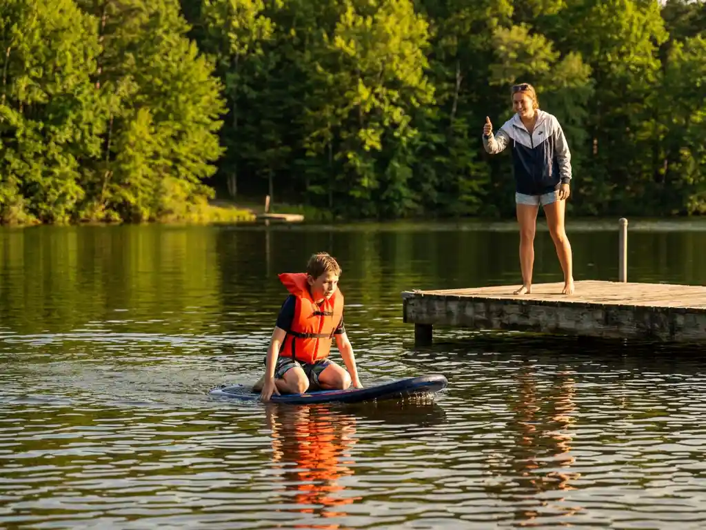 Jonge student in oranje reddingsvest knielt op kneeboard op spiegelend meer, instructeur geeft duim omhoog vanaf houten steiger, groen bos op achtergrond.