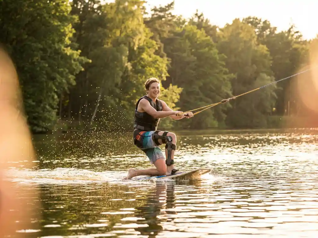 Tiener met waterdichte beenbeugel kneeboardend op een Nederlands meer, gouden zonlicht vangt waterdruppels, groen bos weerspiegelt op het wateroppervlak.