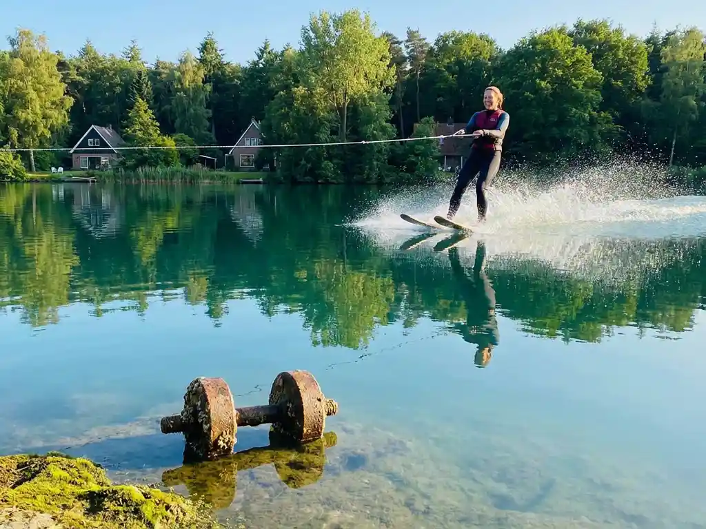 Vrouw waterskiet over turquoise meer met opspattend water, Nederlands bos weerspiegelt in het kalme wateroppervlak, gouden namiddaglicht