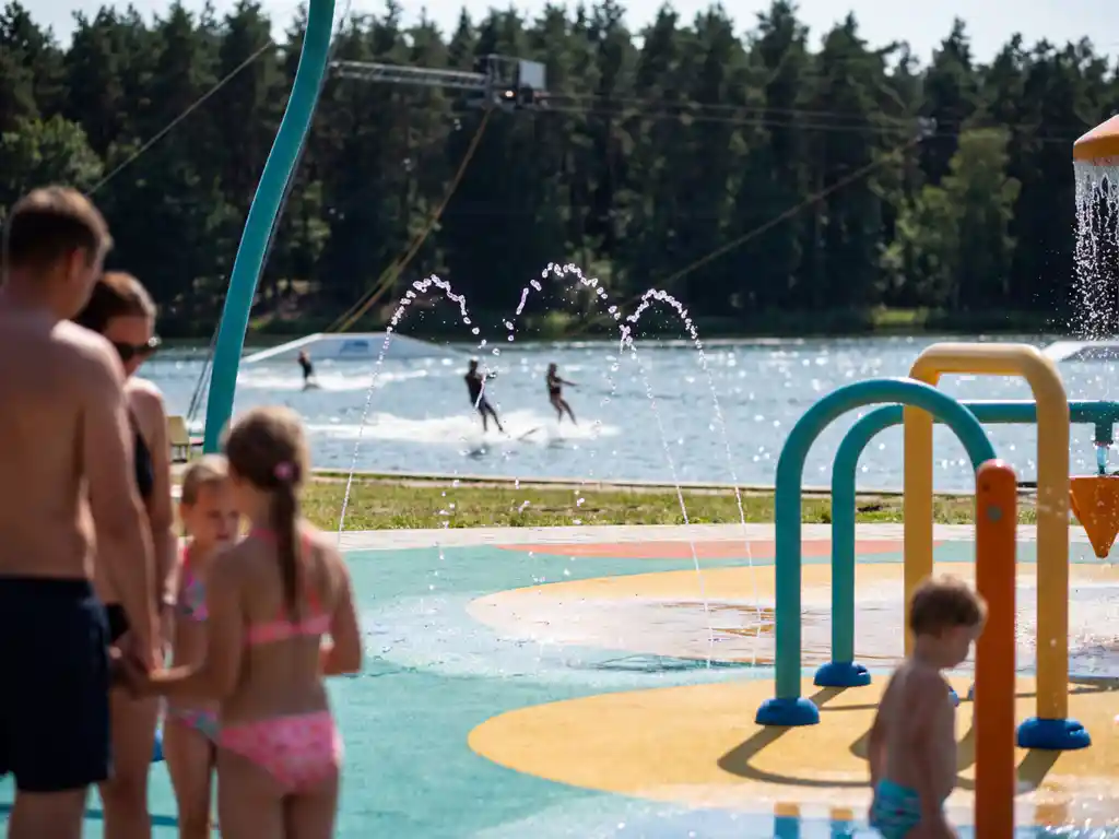 Kinderspeelplaats met fonteinen op de voorgrond, waterskibaan over een meer omgeven door dennenbos op zonnige zomerdag.