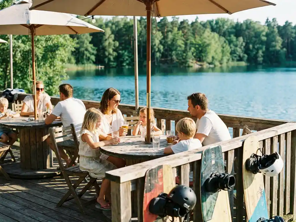 Houten terras aan een blauw meer met wakeboard-uitrusting tegen de reling, gezinnen genieten van drankjes in warm middaglicht