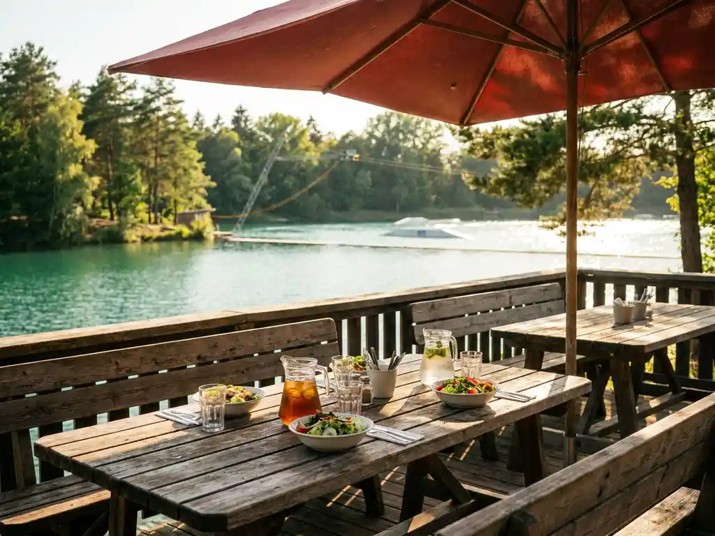 Houten terras met gedekte tafels en rode parasol aan een rustig meer met wakeboard-kabelbaan, omgeven door groene bomen in warm zomerlicht.